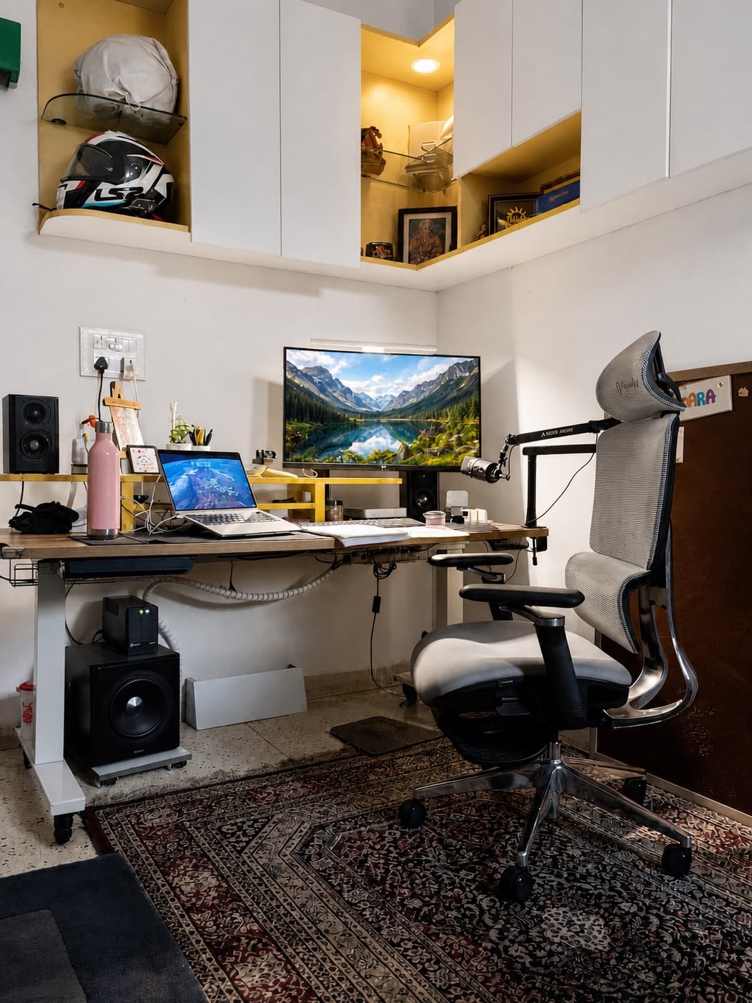Corner standing desk with dual screens, boom microphone, yellow open shelves, and a motorcycle helmet.